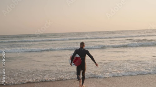 A surfer in a wetsuit runs towards the ocean waves with his surfboard at sunset. He is eager to catch some waves.
