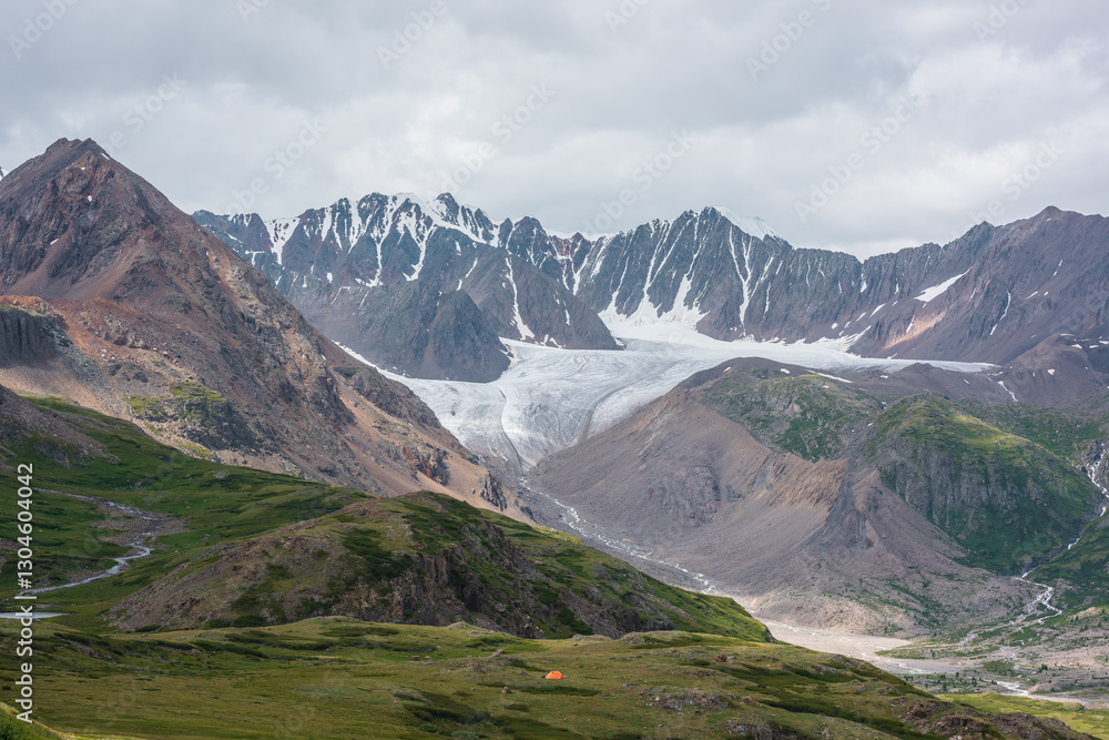 Fototapeta premium Dramatic landscape with orange tent in alpine valley with view to big glacier tongue and large sharp snow-capped mountain range under gray cloudy sky. Green hills and rocks against ice and sheer crags