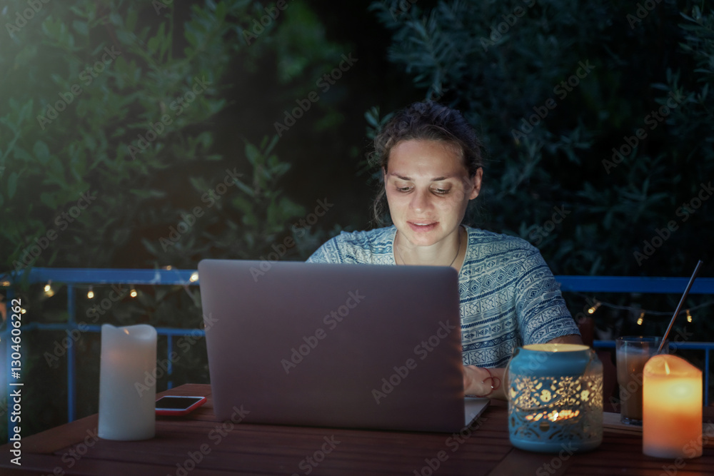 Young hysster woman working on a laptop in the evening on the open terrace of her country house, cozy with candles and lanterns.
