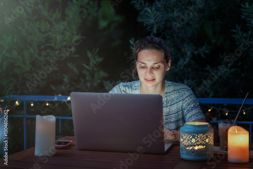 Young hysster woman working on a laptop in the evening on the open terrace of her country house, cozy with candles and lanterns.