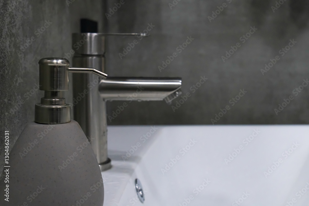 Water dripping from a brushed nickel faucet on a white ceramic sink with grey soap dispenser and concrete wall in background, representing modern bathroom design and hygiene Water dripping from a brushed nickel faucet on a white ceramic sink with grey soap dispenser and concrete wall in background, representing modern bathroom design and hygiene