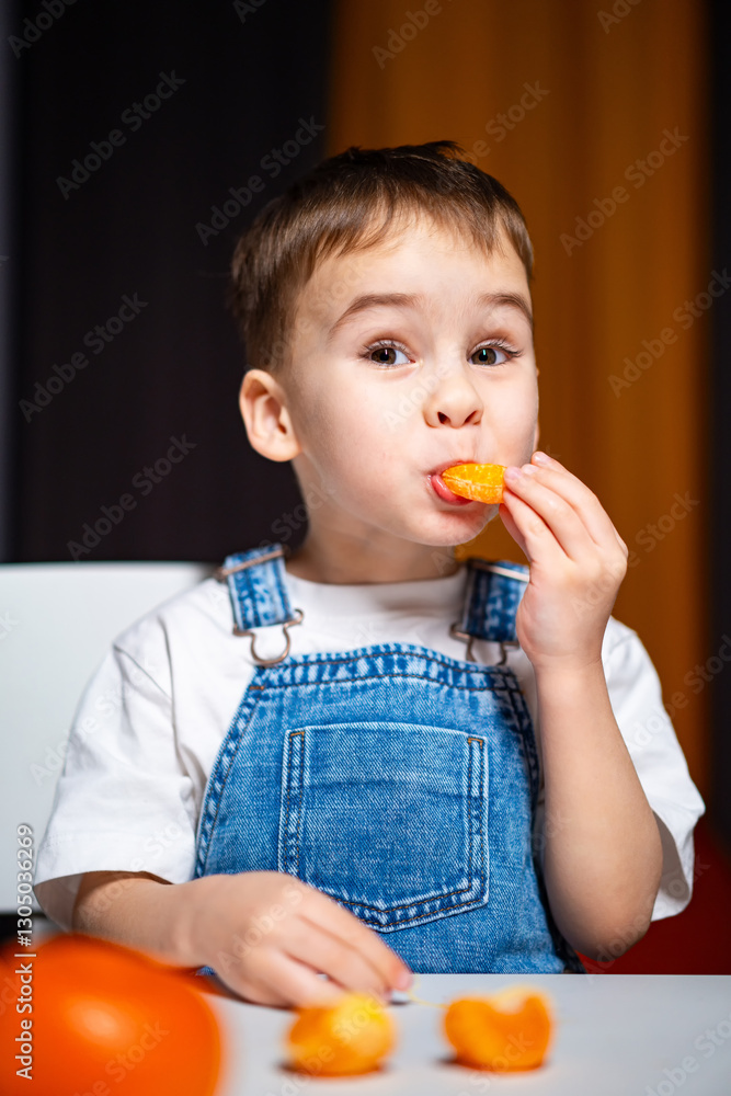Young boy enjoys citrus snack at home. A cheerful young boy in denim overalls tastes a slice of orange, sitting at a table decorated with fruits. Young boy enjoys citrus snack at home. A cheerful young boy in denim overalls tastes a slice of orange, sitting at a table decorated with fruits.