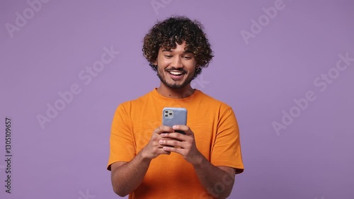 Happy smiling calm young Indian man wear orange t-shirt casual clothes hold use mobile cell phone typing browsing chatting send sms isolated on plain pastel light purple background. Lifestyle concept