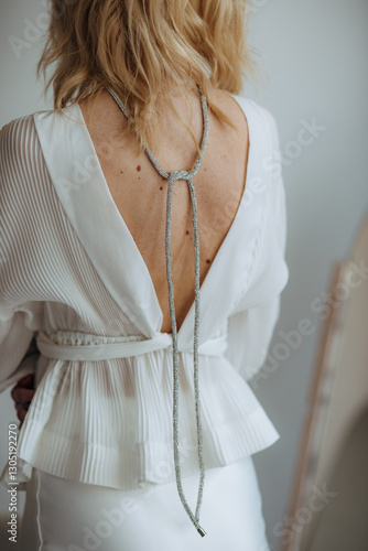a young blonde woman in a white outfit stands with her back bare. on her back is an elegant piece of jewelry. fashionable portrait. Suitable for beauty studios, fashion, cosmetology, jewelry stores 