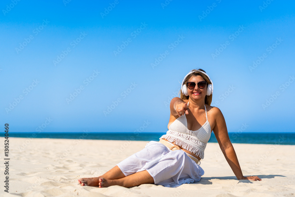 Holidays on beach with music. Beautiful mature woman listening to music in headphones pointing, sitting on white sandy beach. Relaxing in summer Holidays on beach with music. Beautiful mature woman listening to music in headphones pointing, sitting on white sandy beach. Relaxing in summer