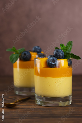 Italian panna cotta dessert of vegan cream thickened with agar agar with layer of mango served in glass garnished with blueberries and mint leaf. Vertical orientation.