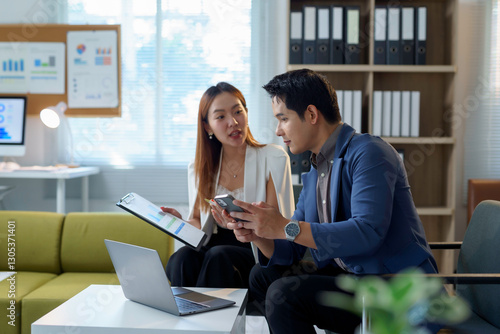Asian business professionals reviewing financial reports, collaborating with laptop and smartphone, analyzing marketing statistics in contemporary workspace