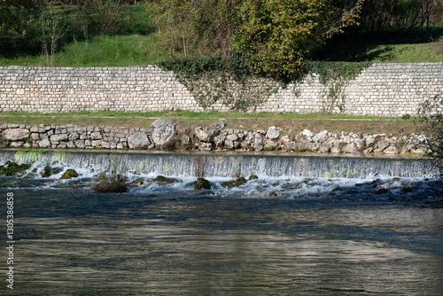 Water falls at the Livenza river resurgences in Polcenigo, Friuli. Picture taken with a telephoto lerns, compressing layers. We see a wooden bench and a stonewall