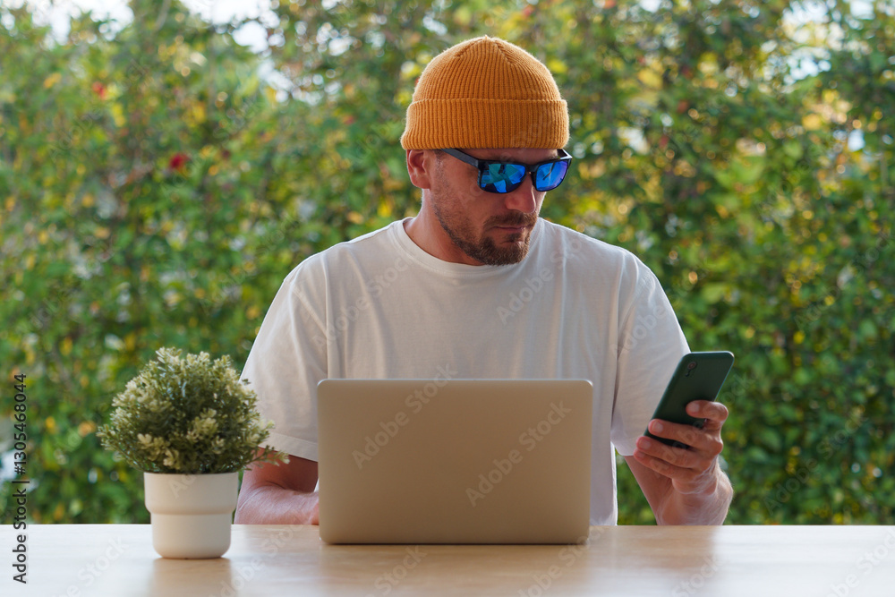 Man dressed in a yellow beanie and sunglasses checks his smartphone while sitting at a wooden table with a laptop. Lush greenery surrounds the outdoor setting. Man dressed in a yellow beanie and sunglasses checks his smartphone while sitting at a wooden table with a laptop. Lush greenery surrounds the outdoor setting.