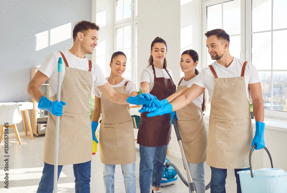 Team of happy cleaners from cleaning service stacking hands. Group of smiling multiethnic janitors in beige aprons and blue rubber gloves standing in office, holding hands together. Teamwork concept Team of happy cleaners from cleaning service stacking hands. Group of smiling multiethnic janitors in beige aprons and blue rubber gloves standing in office, holding hands together. Teamwork concept