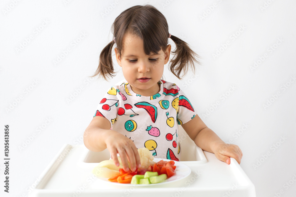 Little girl with pigtails sitting in a high chair and eating fresh vegetables from a white plate, wearing a colorful fruit-patterned t-shirt. Concept of healthy eating, childhood, nutrition Little girl with pigtails sitting in a high chair and eating fresh vegetables from a white plate, wearing a colorful fruit-patterned t-shirt. Concept of healthy eating, childhood, nutrition