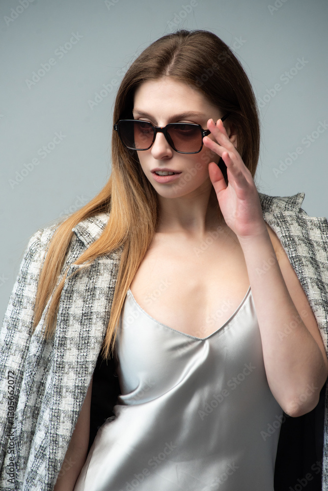 Naklejka premium Elegant woman in a silver satin dress posing stylishly. Accessories include sunglasses and a plaid blazer. Modern, sophisticated, and minimalistic fashion. Neutral background enhances the chic aesthet