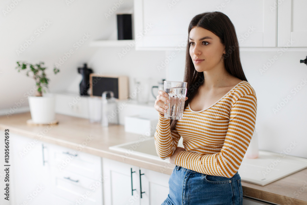 Young European lady holding glass of clean water, looking away, standing in minimalist kitchen interior. Thirst, taking care of health at home and weight loss Young European lady holding glass of clean water, looking away, standing in minimalist kitchen interior. Thirst, taking care of health at home and weight loss