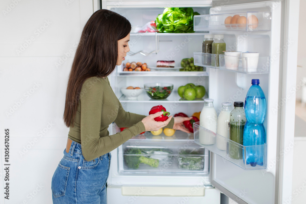Healthy eating concept. Rear view of young woman taking sweet pepper from refrigerator, searching vegetables for preparing meal Healthy eating concept. Rear view of young woman taking sweet pepper from refrigerator, searching vegetables for preparing meal