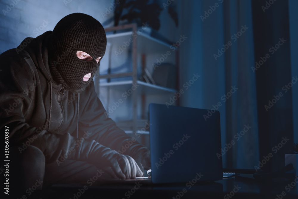 Anonymous Concept. Hacker wearing black balaclava sitting at a desk, downloading private information using laptop at night Anonymous Concept. Hacker wearing black balaclava sitting at a desk, downloading private information using laptop at night
