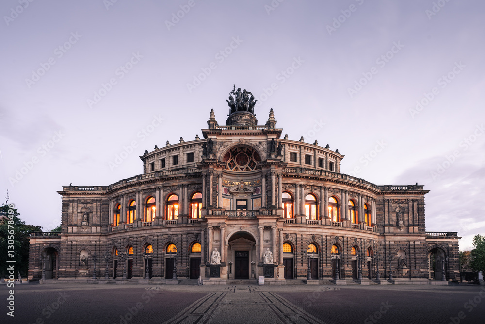 Obraz premium Elegant wide-angle shot of the Semperoper opera house in Dresden, Germany, during twilight. The historic architecture is illuminated by warm interior lights against a soft pastel sky.