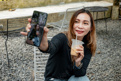 A beautiful woman wearing black clothes and blue jeans showed her cheerful face taking a selfie with the back camera of her cellphone while holding a glass of cold coffee.