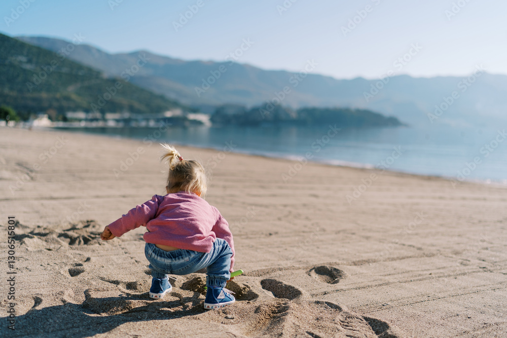Little girl digs sand with a shovel on the beach, squatting down. Back view Little girl digs sand with a shovel on the beach, squatting down. Back view