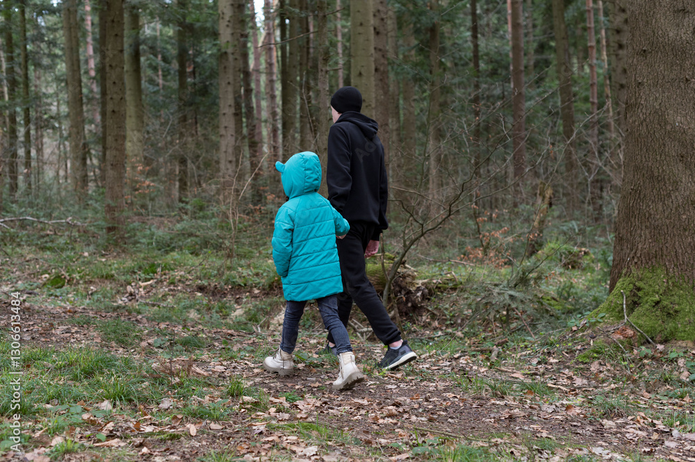 Enjoying a calming walk in the tranquil forest, a child wearing a bright blue jacket strolls alongside an adult in a black hoodie during a cool afternoon Enjoying a calming walk in the tranquil forest, a child wearing a bright blue jacket strolls alongside an adult in a black hoodie during a cool afternoon
