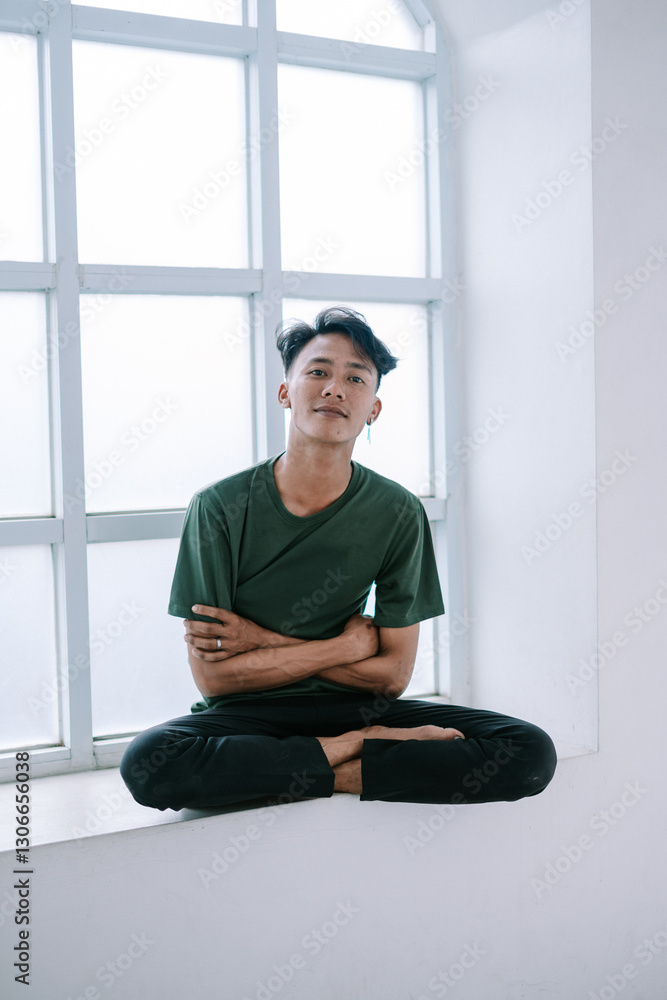 Fototapeta premium Meditative young man sitting near window in relaxed pose indoor studio