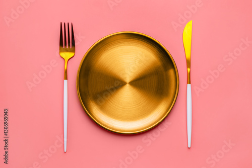 Fototapeta Naklejka Na Ścianę i Meble -  Golden plate with matching fork and knife arranged on a pink background, creating a stylish and elegant table setting for a luxurious dining experience