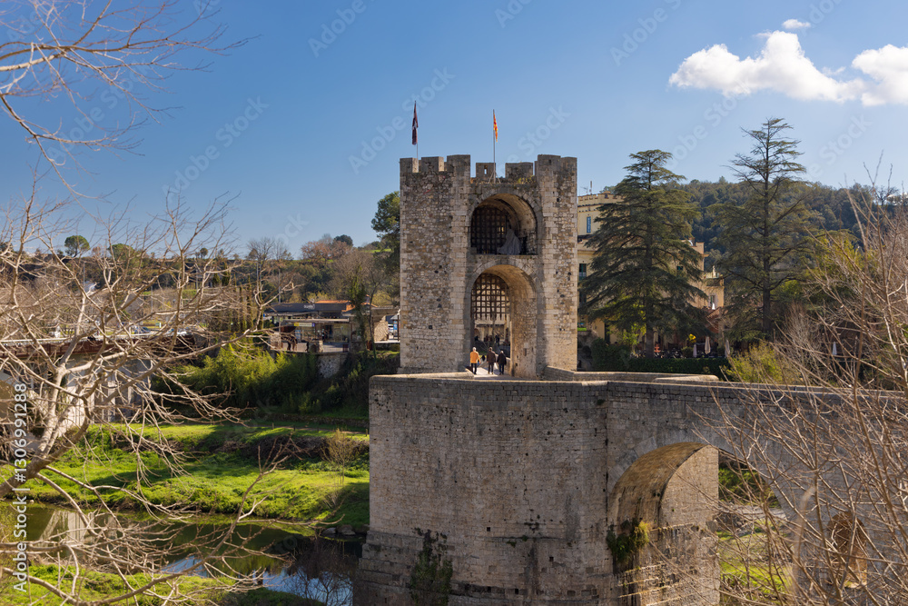 Torre del Pont Vell de Besalú, estructura medieval de piedra con banderas, rodeada de paisaje natural y turistas. Torre del Pont Vell de Besalú, estructura medieval de piedra con banderas, rodeada de paisaje natural y turistas.