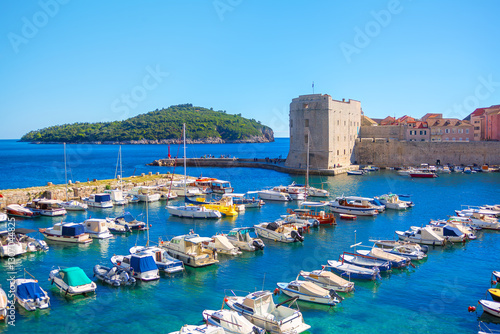 Picturesque view of Dubrovnik, featuring harbor filled with numerous boats and yachts, historic stone tower and city walls. Blue water and green island in distance add to charm of this coastal city