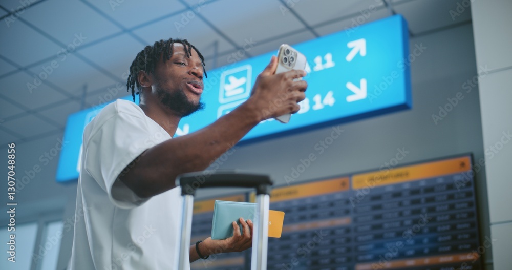 Crowded Airport Terminal: African American Man Talking on Phone by Video Call, Recording Video Content. Arrival and Departure Display, Diverse People Walking in the Background. Tourist Going on Trip. Crowded Airport Terminal: African American Man Talking on Phone by Video Call, Recording Video Content. Arrival and Departure Display, Diverse People Walking in the Background. Tourist Going on Trip.