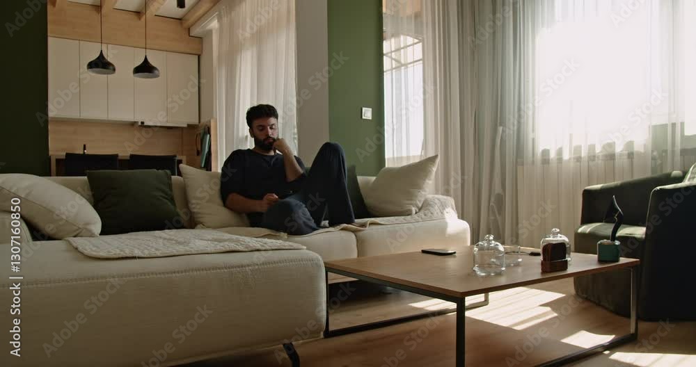 Young man relaxing at home using his cell phone. Caucasian man in his 20s sitting on a couch at home using his cell phone during the day. Young man relaxing at home using his cell phone. Caucasian man in his 20s sitting on a couch at home using his cell phone during the day.