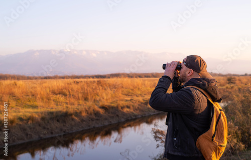A man in a cap and with a yellow backpack on a sunny day in spring in the thickets of bushes watches birds and wild animals with binoculars. Bird watching