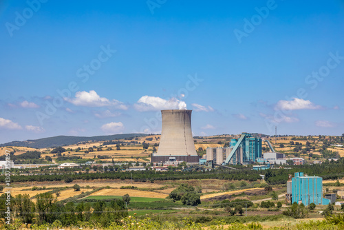 A thermal power plant in Çan, Çanakkale, Turkey, surrounded by agricultural fields and rolling hills. The industrial facility contrasts with the natural landscape under a clear blue sky.