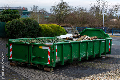 A green dumpster is located in an urban setting, filled with various construction debris and materials. Surrounding foliage is visible, indicating the presence of nearby buildings and infrastructure.