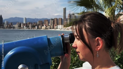 Tourist woman observing expansive Benidorm cityscape and mediterranean coastline through stationary coin-operated telescope at sunny outdoor viewpoint with coastal panorama. Close-up