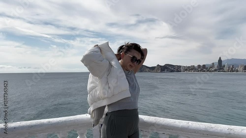 Tourist woman wears a white jacket observing expansive Benidorm cityscape and mediterranean coastline at sunny outdoor viewpoint with coastal panorama. Medium shot