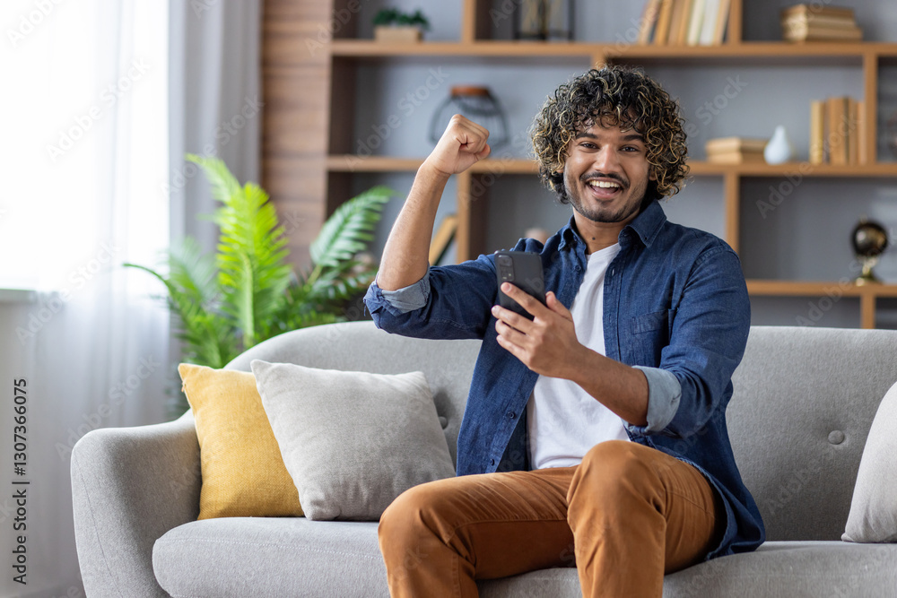Obraz premium Cheerful man with curly hair celebrating success while using a smartphone at home, sitting comfortably on the sofa.