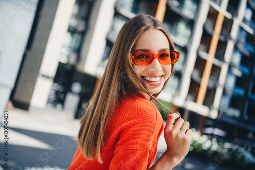 Young woman in stylish orange shirt and sunglasses, smiling while walking in urban setting during a sunny day photo