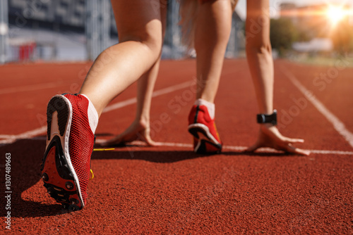 Fototapeta Naklejka Na Ścianę i Meble -  Female athlete at the starting line of a sprint race, wearing red track spikes, ready for competition on a sunlit outdoor stadium track.