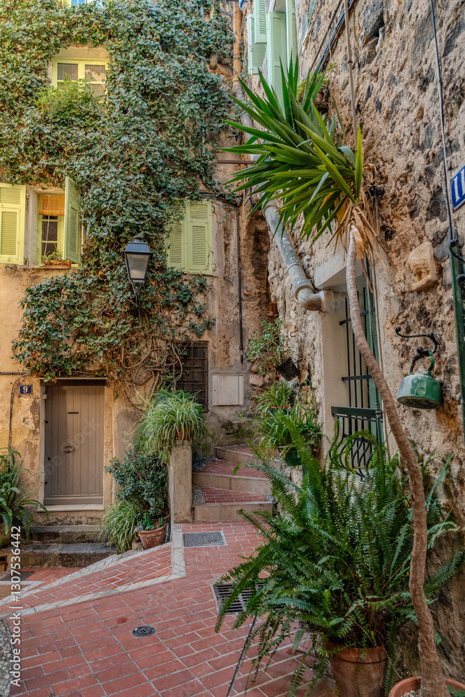 Naklejka premium Charming Old Town Street In Menton: Narrow Cobblestone Alley With Colorful Mediterranean Buildings, Green Wooden Shutters, Hanging Laundry, Potted Plants, And Aged Facades In The French Riviera