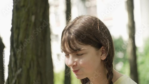 A teenage girl with pigtails in a yellow t-shirt is making funny faces and laughing while chewing food in a city park on a sunny summer day. Left-side shot, close-up