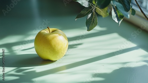 A golden delicious apple shines in soft light beside leafy branches on a tranquil, green surface.
