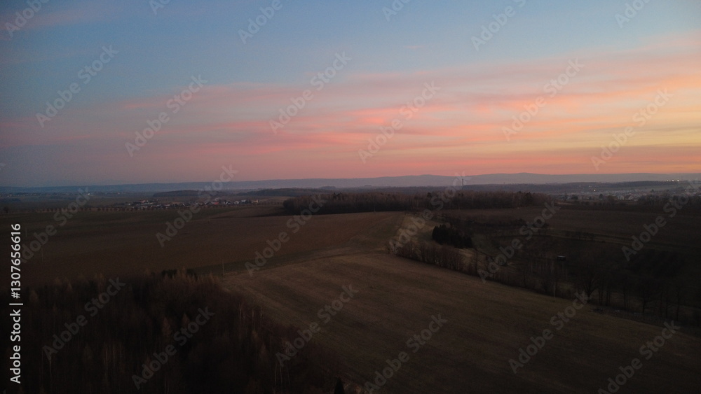 Obraz premium The dark silhouette of the landscape contrasts beautifully against the colorful clouds. In the background, rolling hills stretch across the horizon, with a wind turbine visible in the distance.