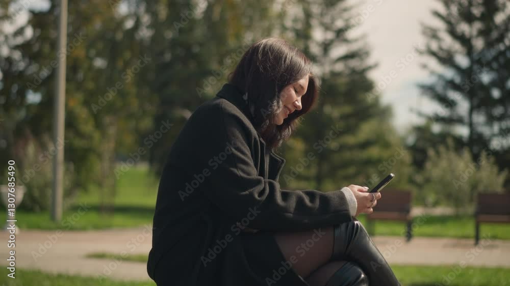 Side view of professional woman with leg crossed, happily focused on phone, blur chair in a park setting with greenery around, sunny day Side view of professional woman with leg crossed, happily focused on phone, blur chair in a park setting with greenery around, sunny day