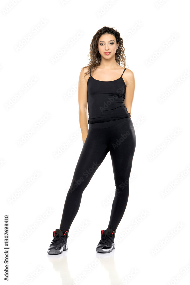 Full-length portrait of young woman with wavy hair wearing black tank top, leggings and athletic shoes, standing confidently against white background Full-length portrait of young woman with wavy hair wearing black tank top, leggings and athletic shoes, standing confidently against white background