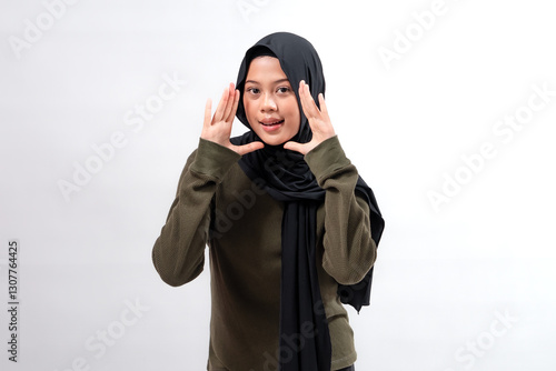 Indonesian Muslim woman wearing a black hijab and olive sweater, smiling while making a calling gesture with both hands. Studio portrait with soft lighting against a white background.