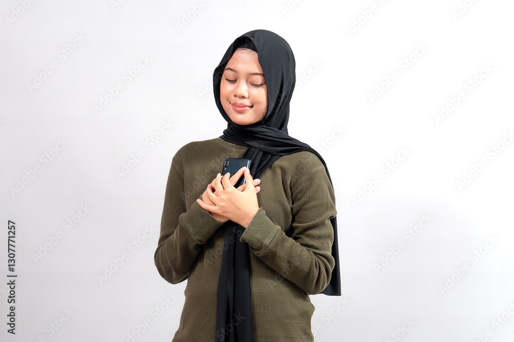 A young Indonesian woman in a black hijab and olive-green sweater holds a smartphone close to her heart with a peaceful smile. She stands against a plain white background, radiating warmth. A young Indonesian woman in a black hijab and olive-green sweater holds a smartphone close to her heart with a peaceful smile. She stands against a plain white background, radiating warmth.