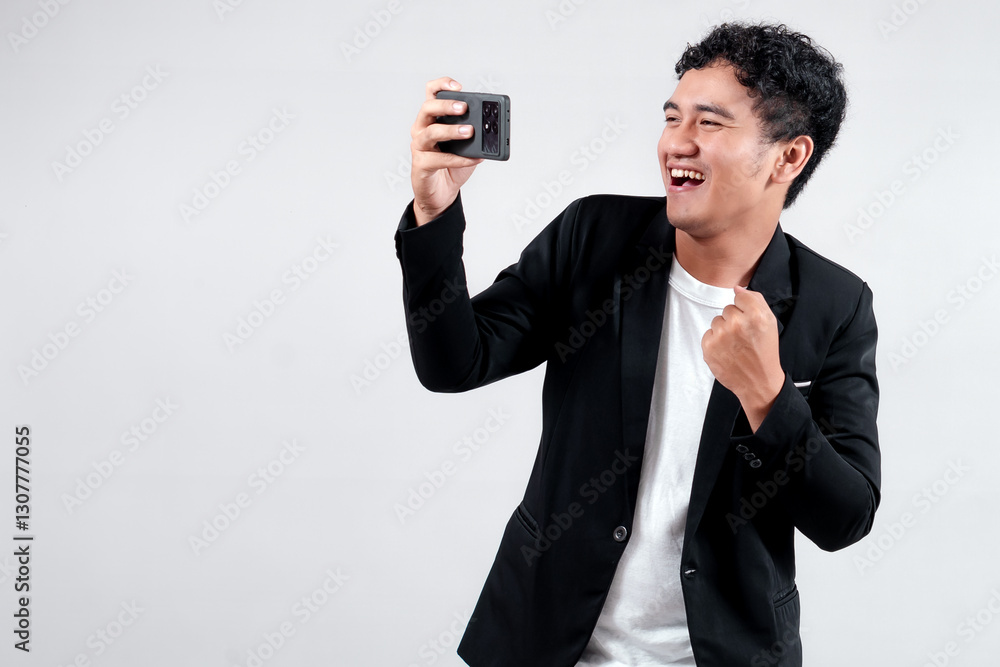 Asian man in a black blazer and white shirt celebrating victory while looking at his smartphone. He is smiling with excitement, raising his fist joyfully in a success gesture. Asian man in a black blazer and white shirt celebrating victory while looking at his smartphone. He is smiling with excitement, raising his fist joyfully in a success gesture.