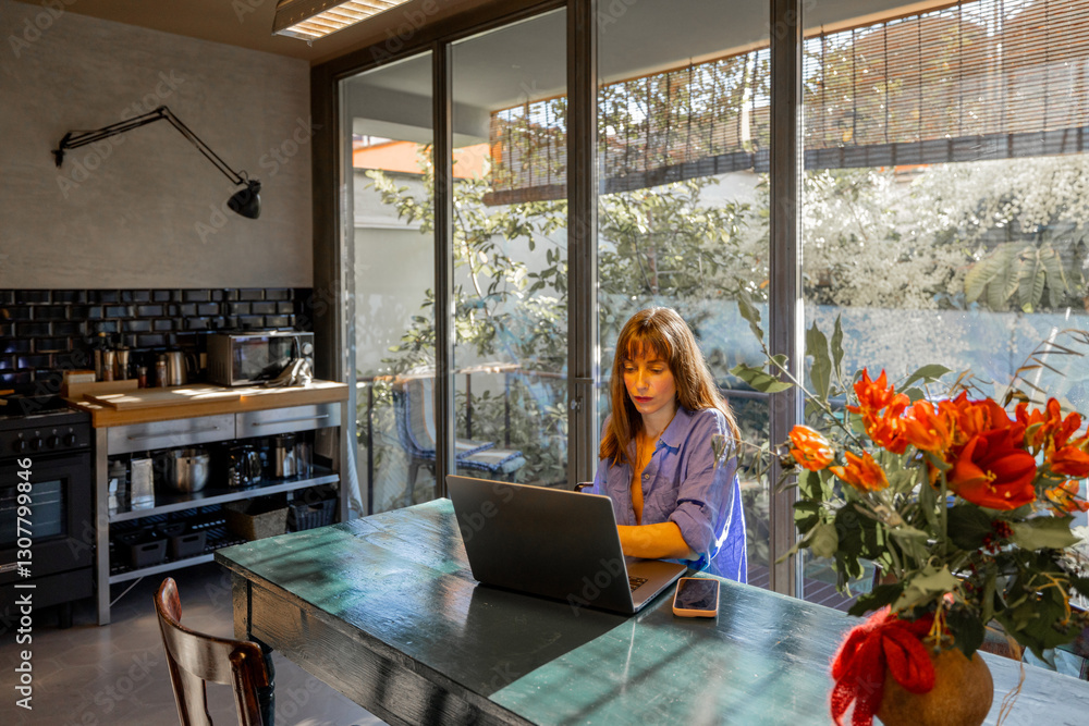 A woman in a relaxed blue outfit works on her laptop at a rustic dining table in a modern kitchen. Large glass windows fill the space with natural light, creating a serene and productive atmosphere