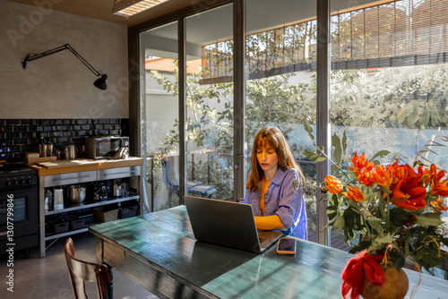 Obraz na płótnie A woman in a relaxed blue outfit works on her laptop at a rustic dining table in a modern kitchen. Large glass windows fill the space with natural light, creating a serene and productive atmosphere w salonie