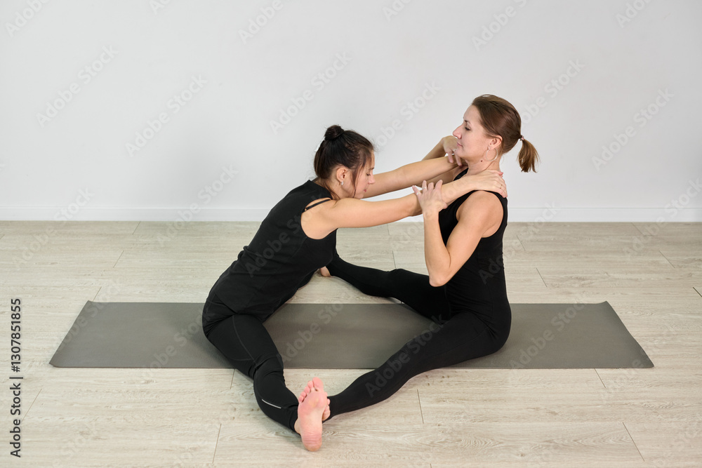 Obraz premium Young adult women engaging in partner yoga on wooden floor, one assisting other in stretch. Setting is calm, with minimal decor and natural lighting, evoking tranquil mood