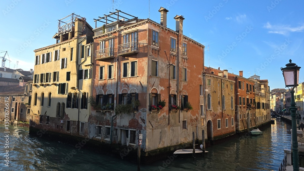 Fototapeta premium A weathered Venetian building with peeling facades and elegant balconies sits at the intersection of two canals. Small boats float nearby, and flower-filled windows add charm to the historic waterfron